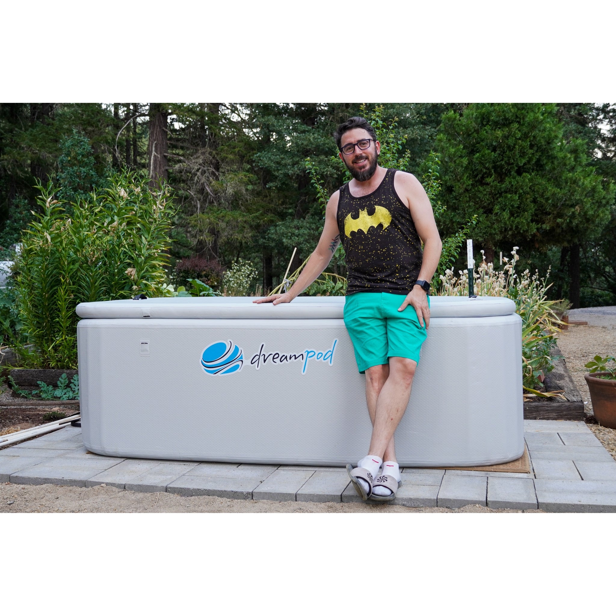 Man leaning against a Dreampod rectangular ice bath with beige exterior and logo, installed on a paved patio surrounded by trees and greenery