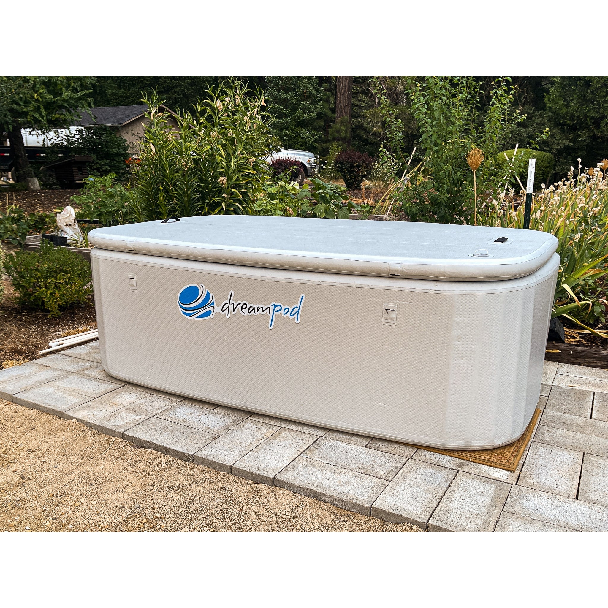Dreampod rectangular ice bath with beige exterior and logo, installed on a paved patio surrounded by trees and garden greenery