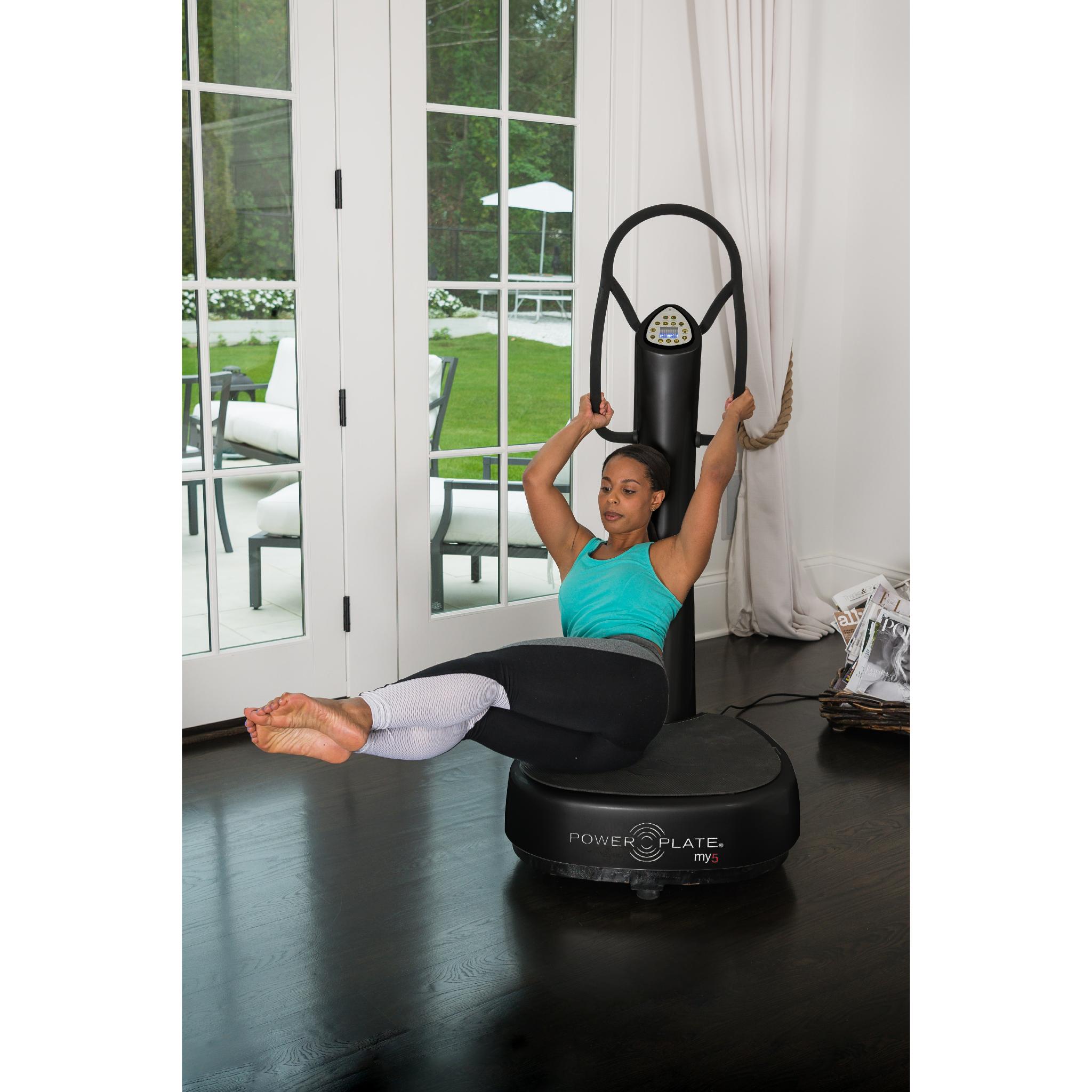Woman performing a raised leg core exercise on a black Power Plate my5 vibration plate with straight handlebars in a bright home setting near large glass doors