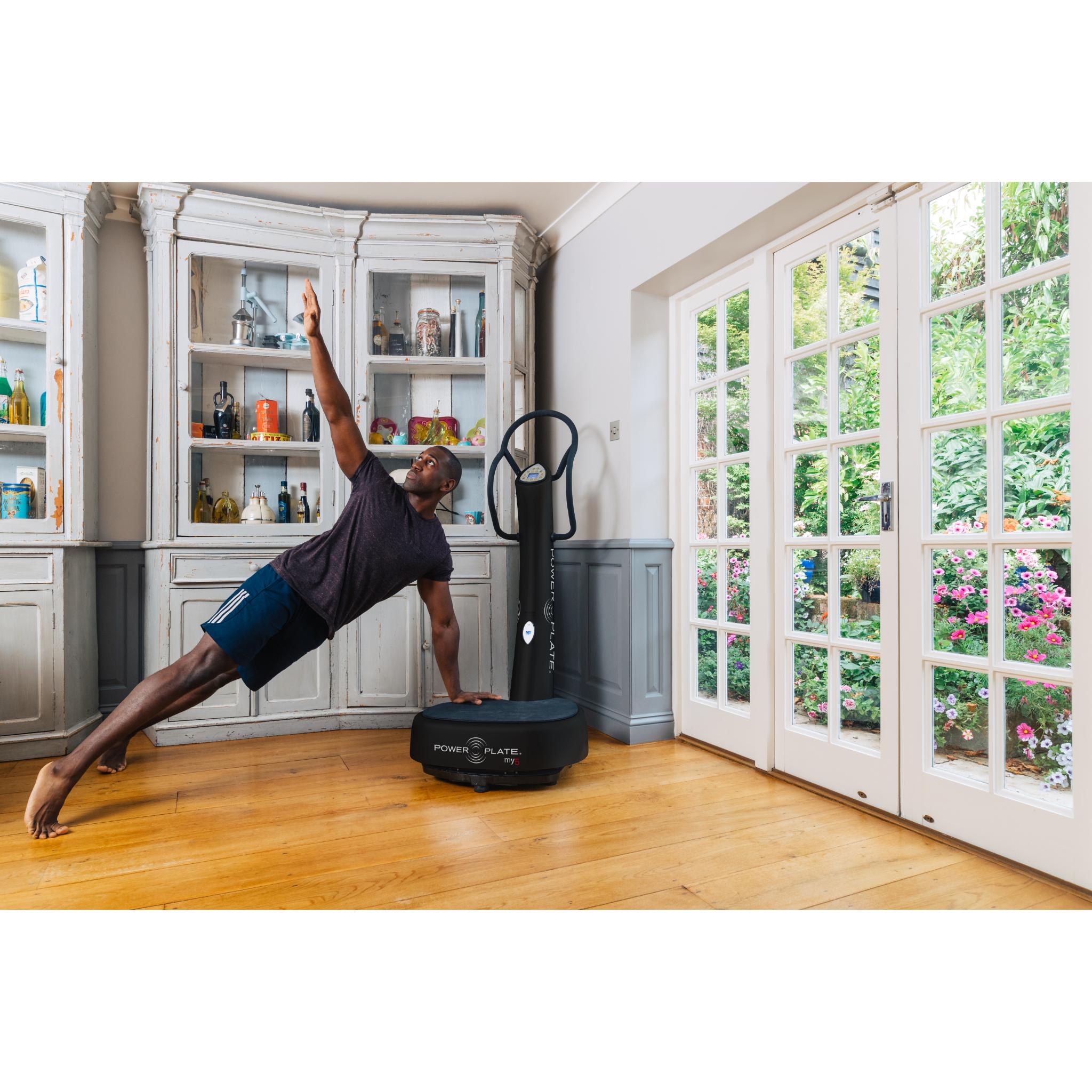 Man performing a side plank on a black Power Plate my5 vibration plate with straight handlebars in a bright home setting near large glass doors