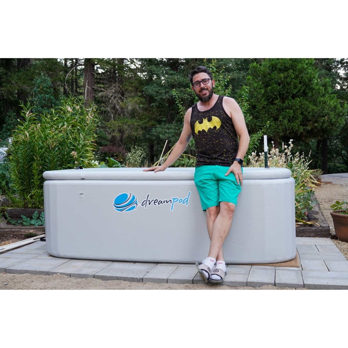 Man leaning against a Dreampod rectangular ice bath with beige exterior and logo, installed on a paved patio surrounded by trees and greenery