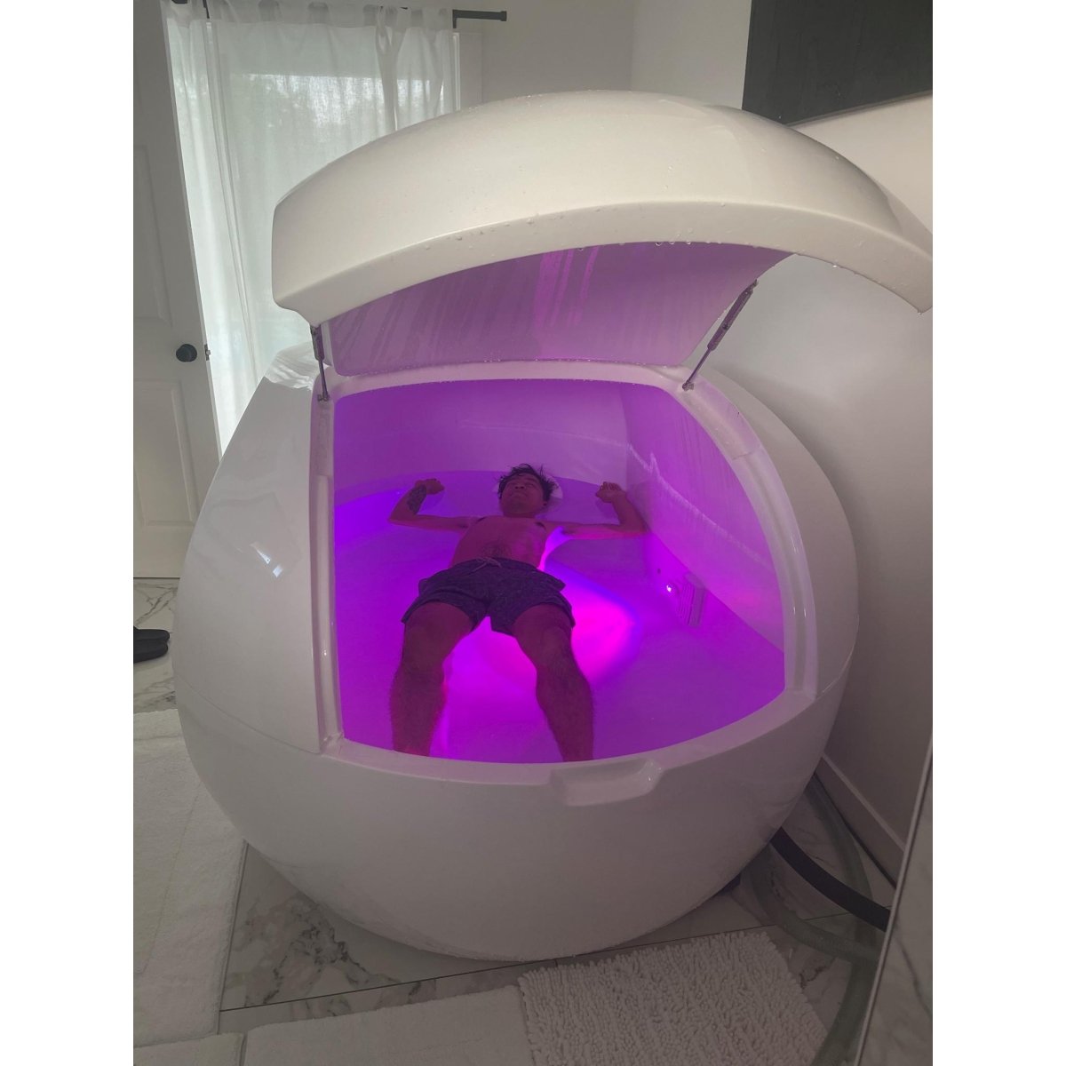 Modern white float pod with canopy lid, person inside illuminated by purple LED lighting for relaxation and sensory therapy