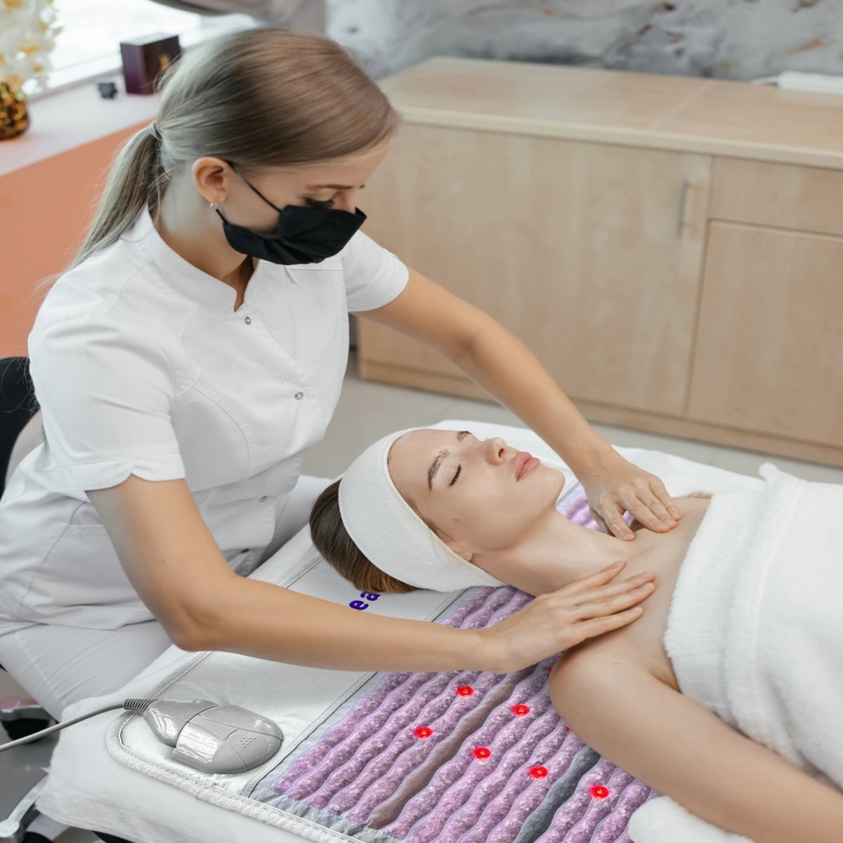 Spa professional providing facial treatment while client relaxes on HealthyLine Platinum Aura Mat Pro Plus 7428 with gemstone therapy