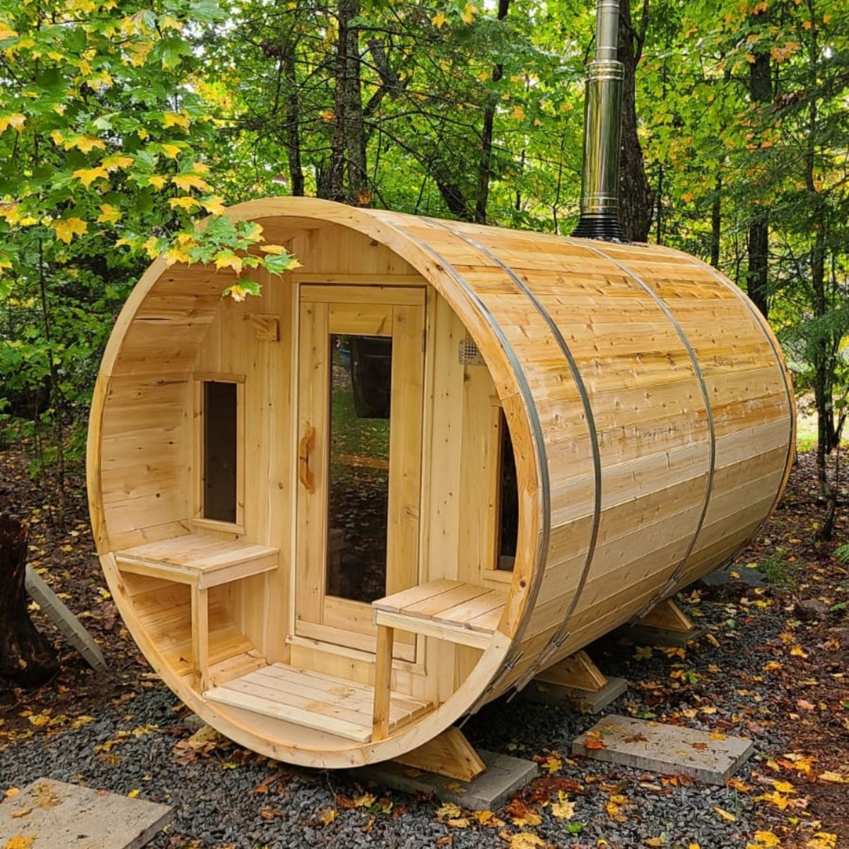 Leisurecraft CT Tranquility Barrel Sauna in natural cedar wood finish, placed outdoors in a lush forest with open glass door and benches inside, showcasing craftsmanship and inviting rustic design.
