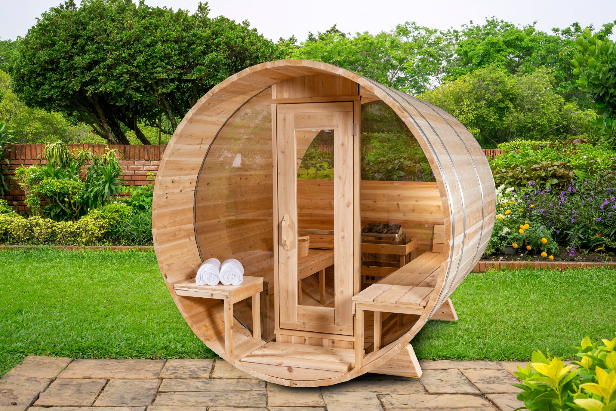 Leisurecraft Serenity MP Barrel Sauna with open glass door revealing cedar benches, set on a paved patio surrounded by lush green garden scenery.