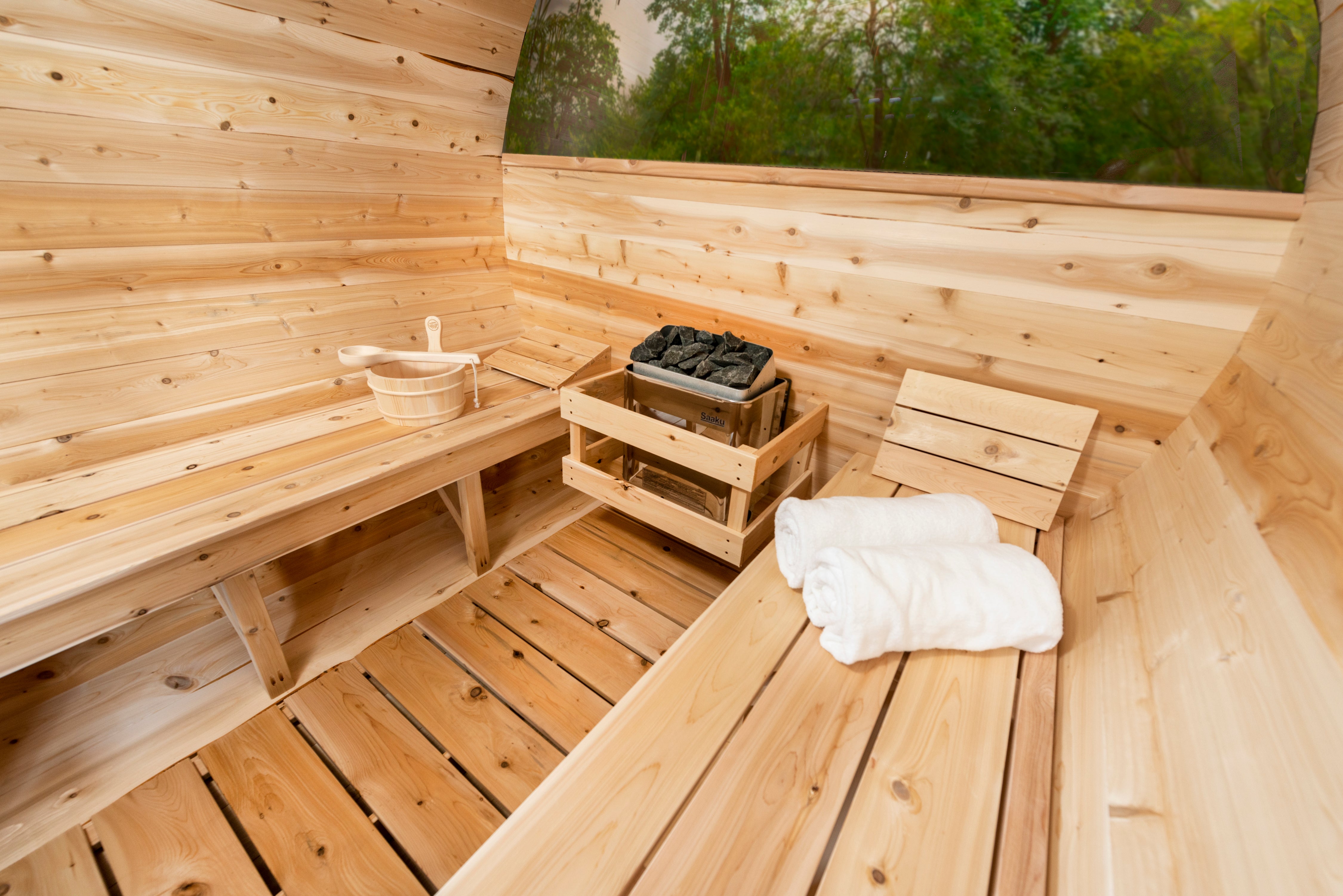 Leisurecraft Serenity MP Barrel Sauna interior featuring smooth cedar benches, wooden basket, neatly folded towels, and a traditional sauna heater with stones.