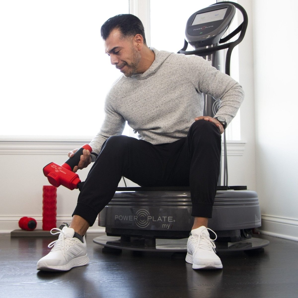 Man using Power Plate Pulse massage gun on legs while sitting on Power Plate machine