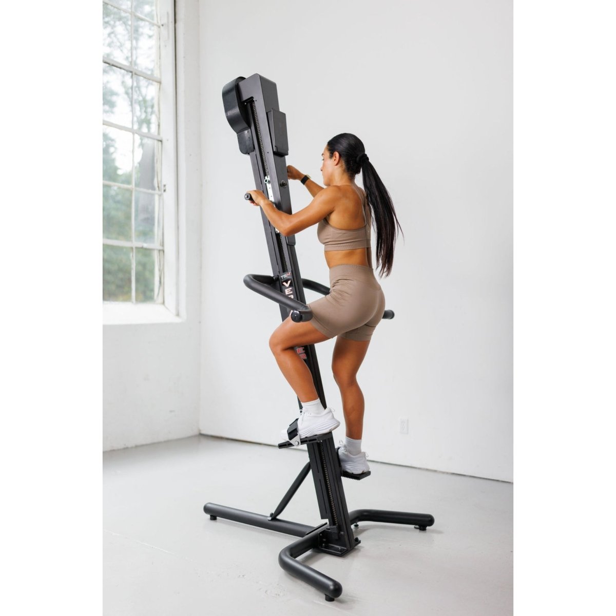 Woman exercising on the VersaClimber TS-M Magnetic in regular black, demonstrating full-body zero-impact cardio and strength training in a bright studio with large windows.