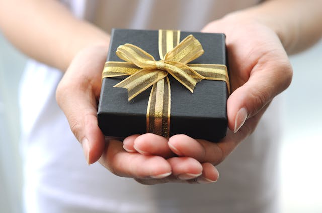 Black gift box with a gold ribbon held by two hands against a neutral background