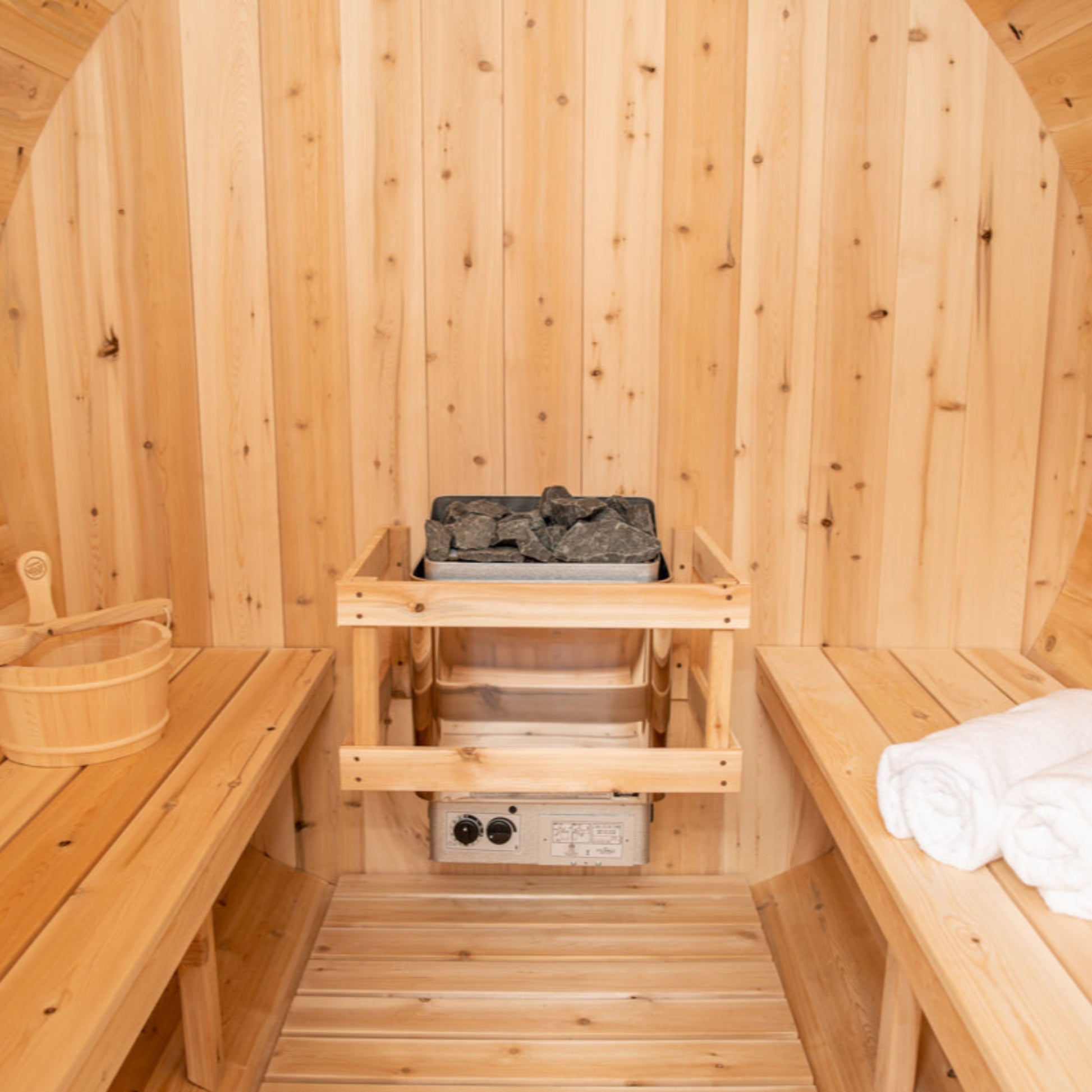 Interior of a cedar barrel sauna featuring wooden benches, rolled towels, and an electric heater with stones.