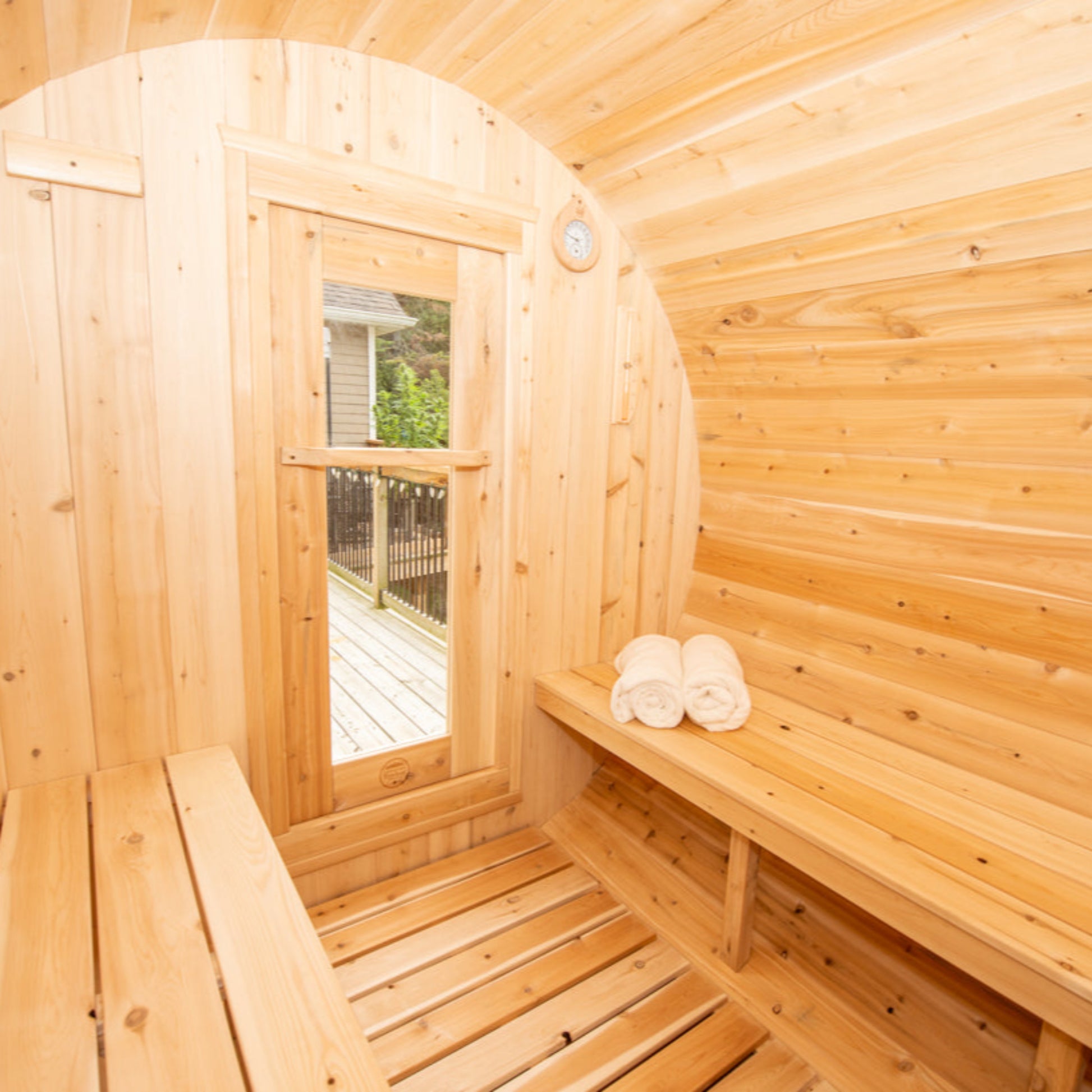 Interior of a cedar barrel sauna featuring wooden benches, a side window with outdoor deck view, and neatly rolled towels.