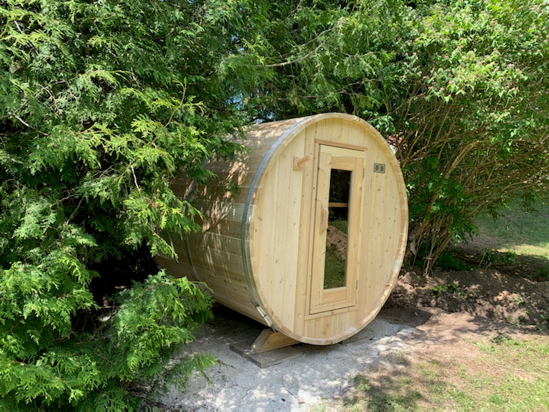 Outdoor cedar barrel sauna nestled in a lush garden, surrounded by greenery and shaded by tall trees.