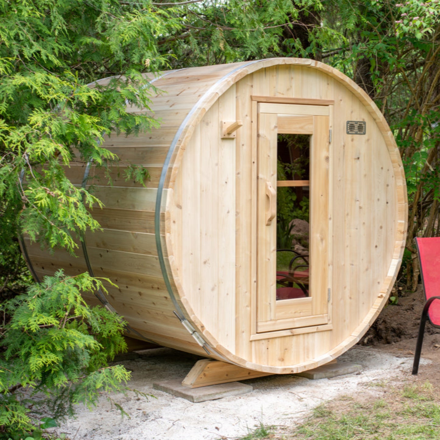 Outdoor cedar barrel sauna placed in a natural garden setting, surrounded by greenery, with a wooden door and curved roof.