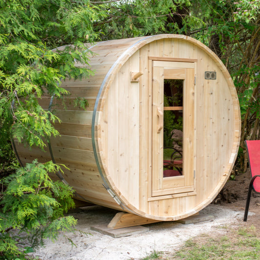 Outdoor cedar barrel sauna placed in a natural garden setting, surrounded by greenery, with a wooden door and curved roof.