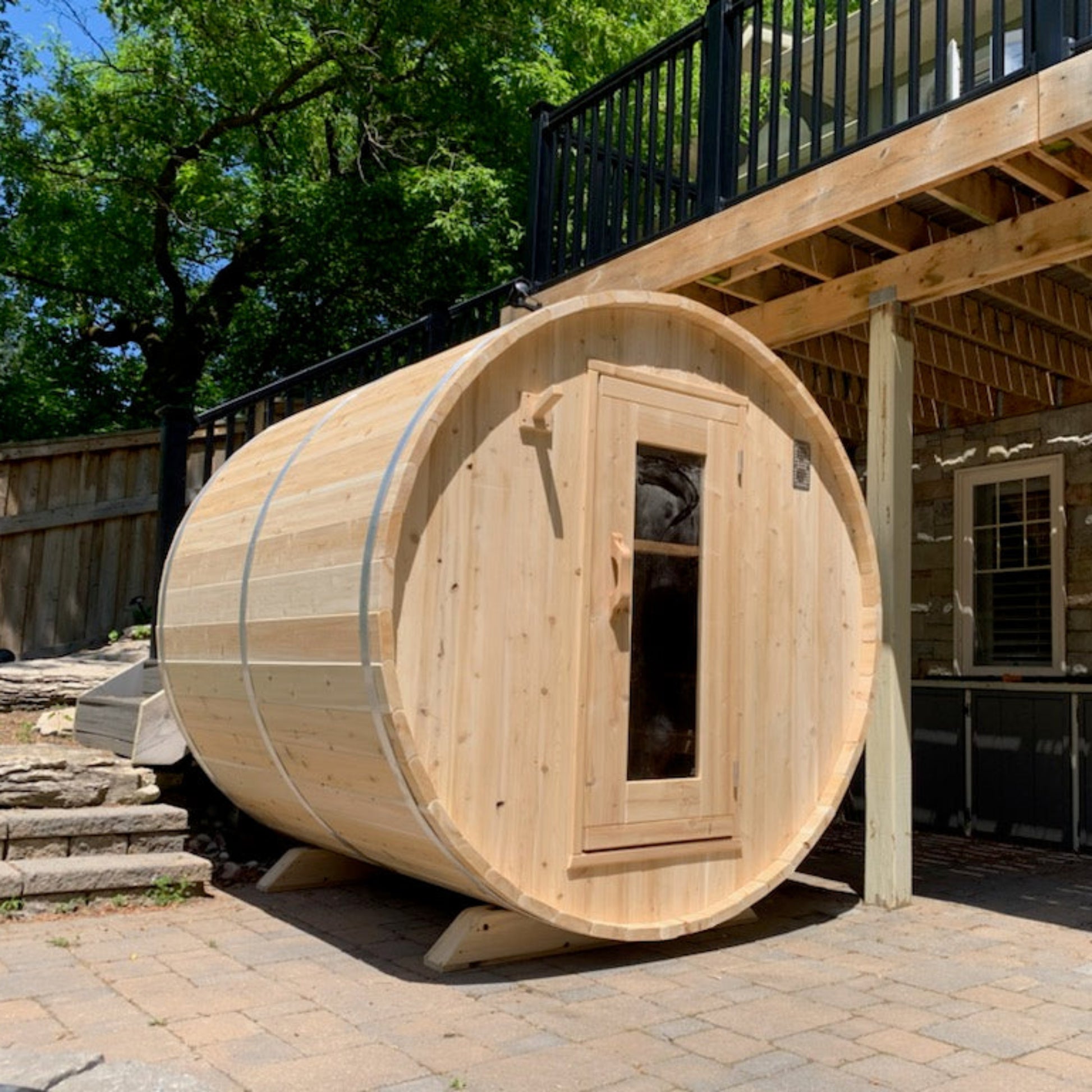 Outdoor cedar barrel sauna set on a stone patio beneath a wooden deck, framed by greenery and shaded by tall trees.