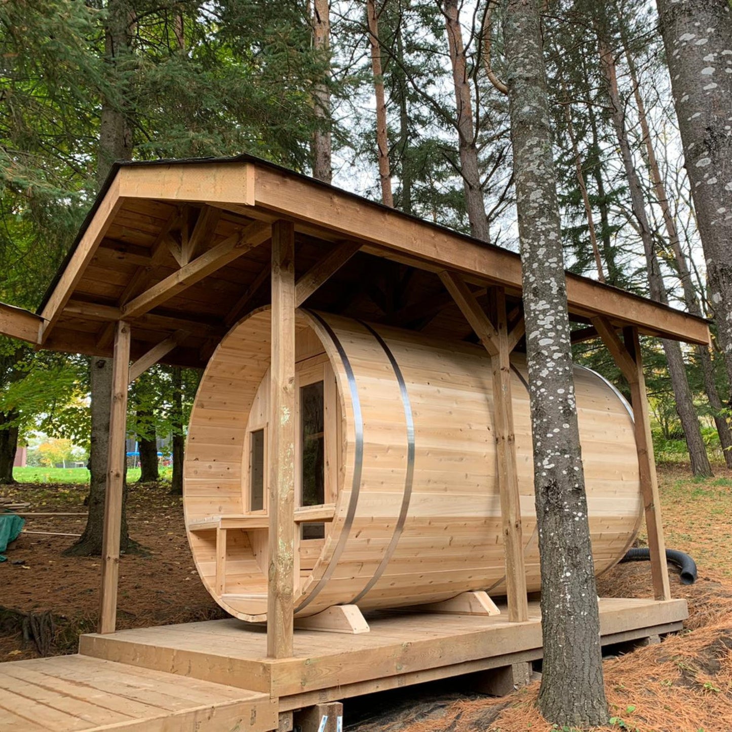 Leisurecraft Canadian Timber barrel sauna in natural wood finish, displayed outdoors with extended porch roof, glass door, and benches inside, highlighting durable craftsmanship