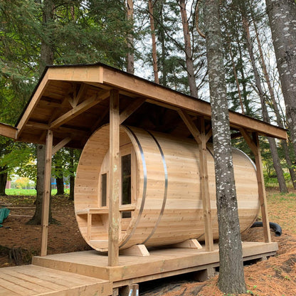 Leisurecraft Canadian Timber barrel sauna in natural wood finish, displayed outdoors with extended porch roof, glass door, and benches inside, highlighting durable craftsmanship