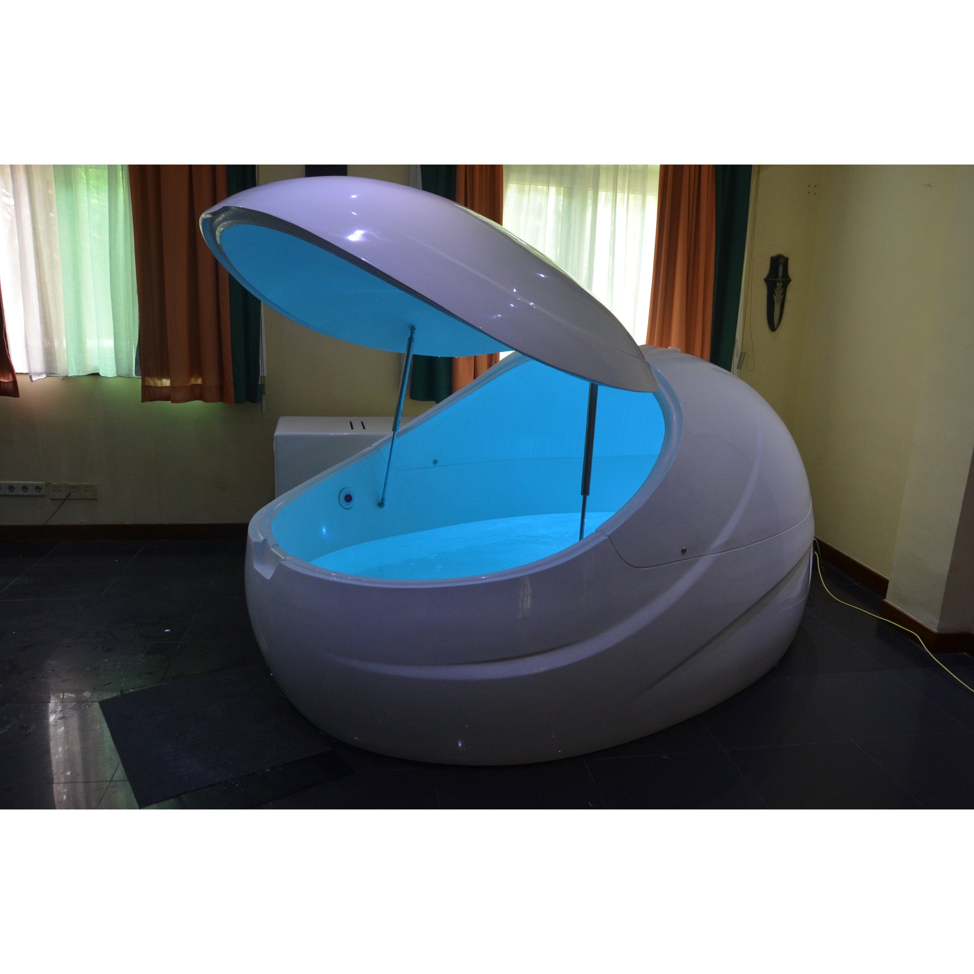 Dreampod float pod with sleek white finish and open canopy lid, illuminated with calming blue LED lighting, placed in a cozy wellness room with curtains for relaxation therapy