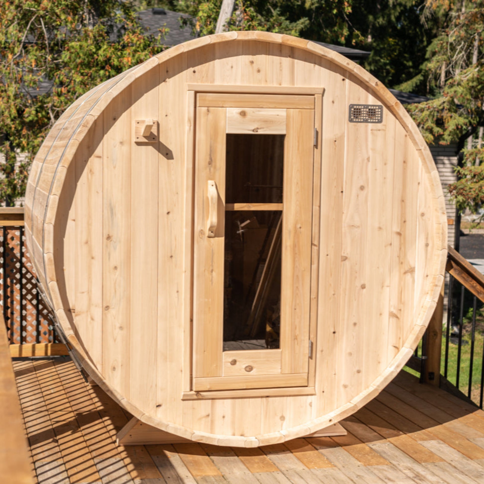 Outdoor cedar barrel sauna on a raised wooden deck with trees in the background, featuring a front glass door and rounded shape.