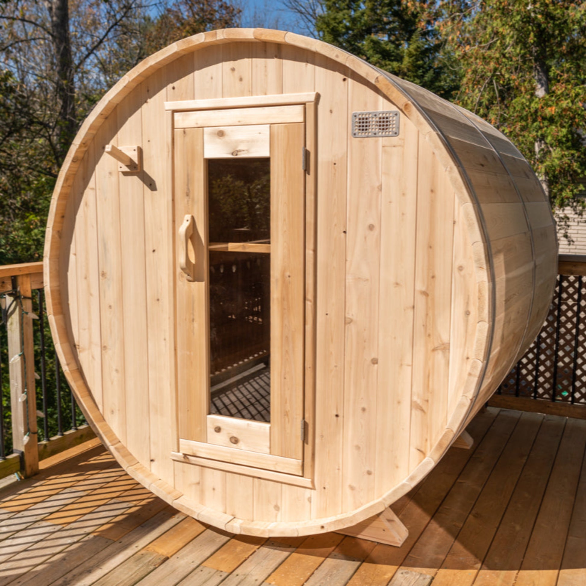 Outdoor cedar barrel sauna on a raised wooden deck with trees in the background, featuring a front glass door and rounded barrel design.