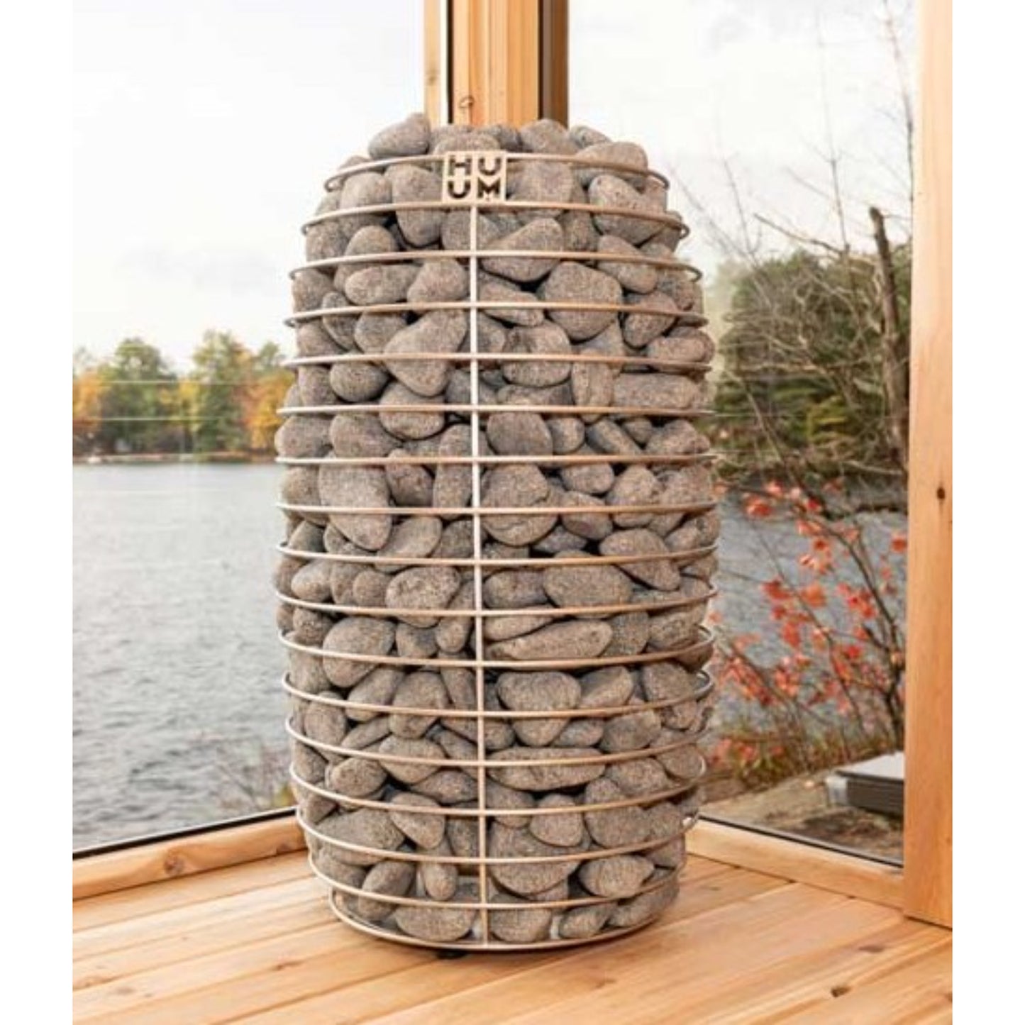 Tall HUUM HIVE sauna heater filled with natural stones, placed on a wooden deck with a lake view in the background
