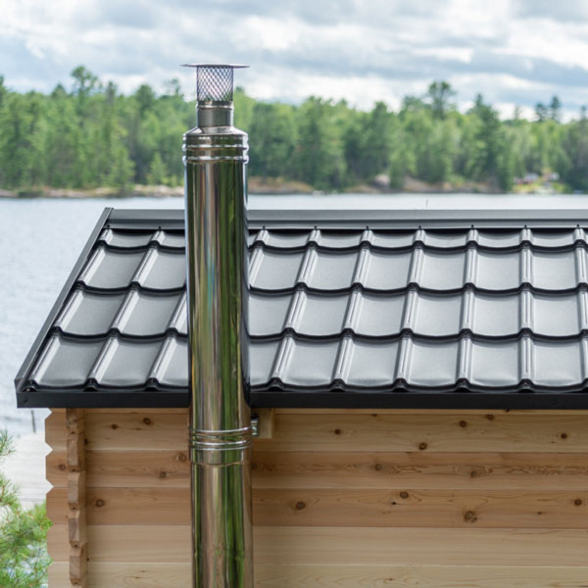Barrel sauna with a black metal roof and stainless steel chimney pipe, set outdoors near a lake with trees in the background
