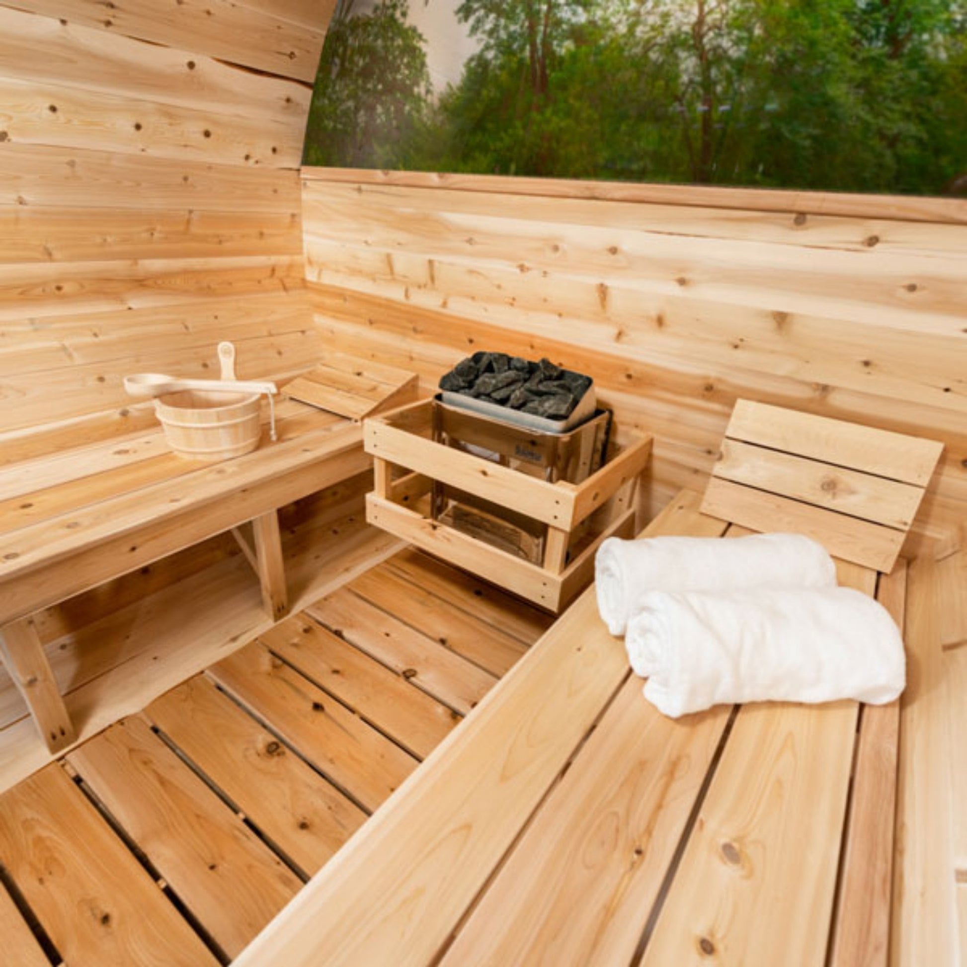 Wooden barrel sauna interior showing cedar benches, sauna heater with stones, wooden water bucket, and towel