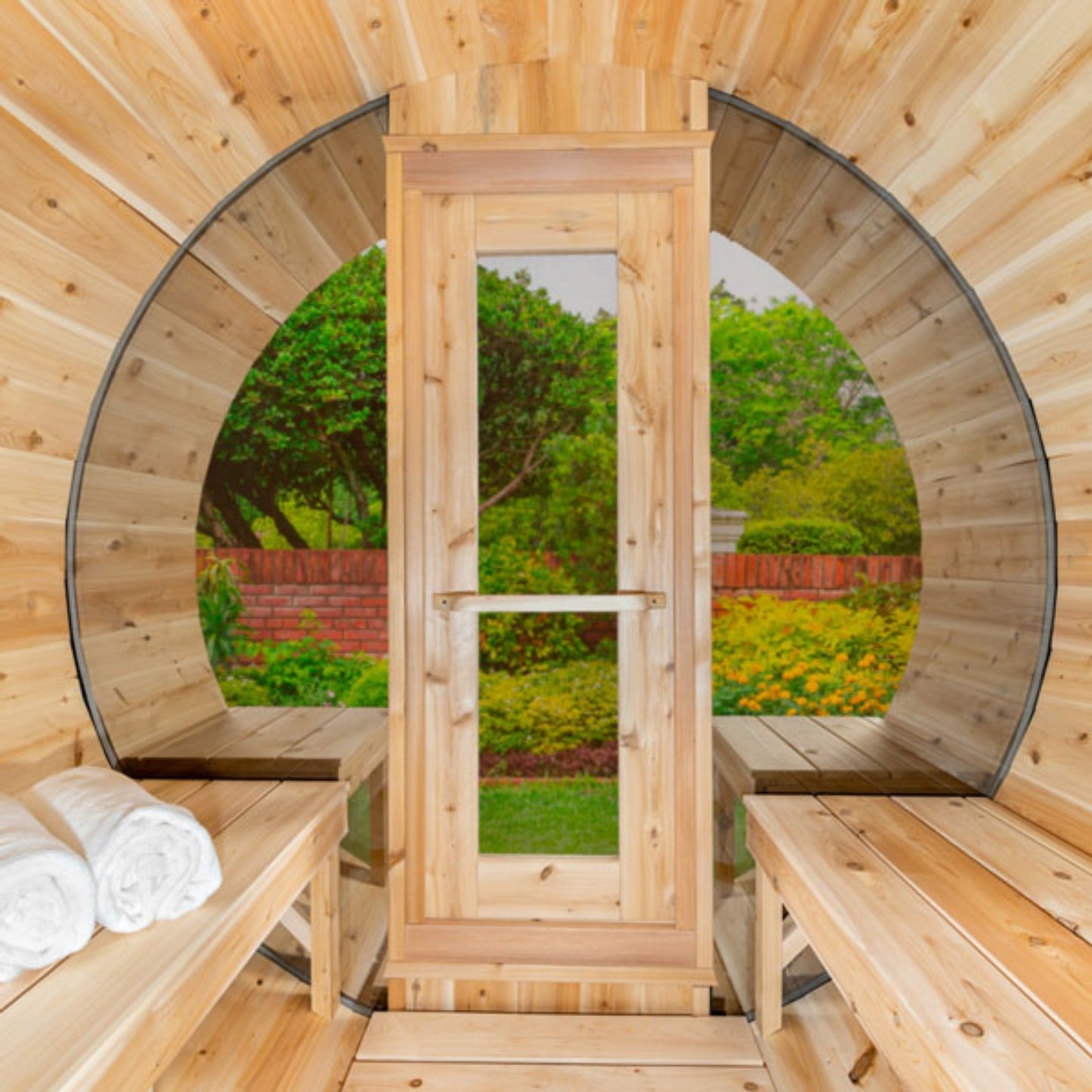 Inside view of wooden barrel sauna with benches and towels, facing the front glass door open to a garden