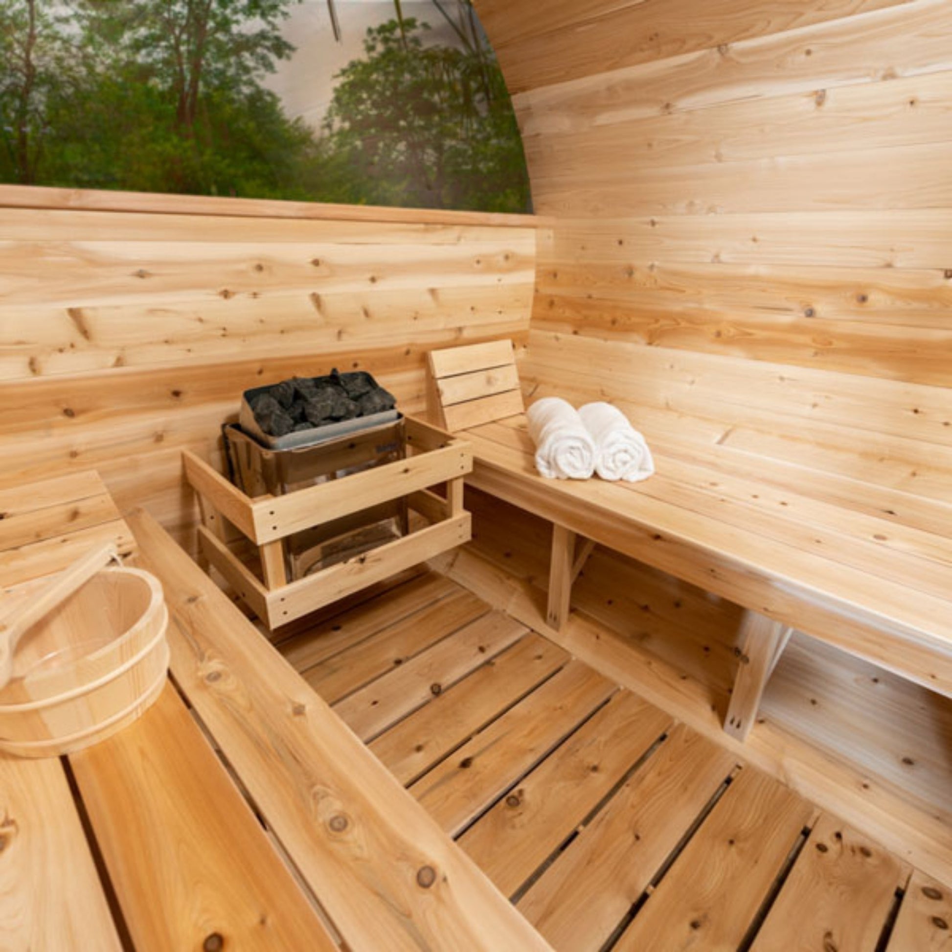 Wooden barrel sauna interior with cedar benches, sauna heater, towel, and wooden water bucket on the floor