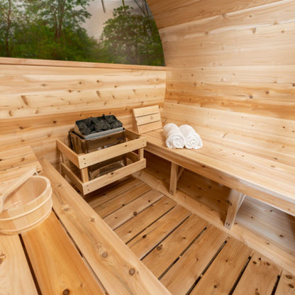 Wooden barrel sauna interior with cedar benches, sauna heater, towel, and wooden water bucket on the floor