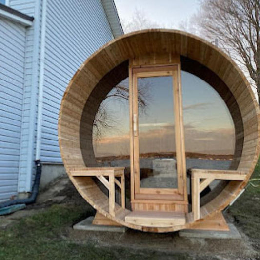 Wooden barrel-shaped outdoor sauna with front door, windows, and small porch seating placed beside a building