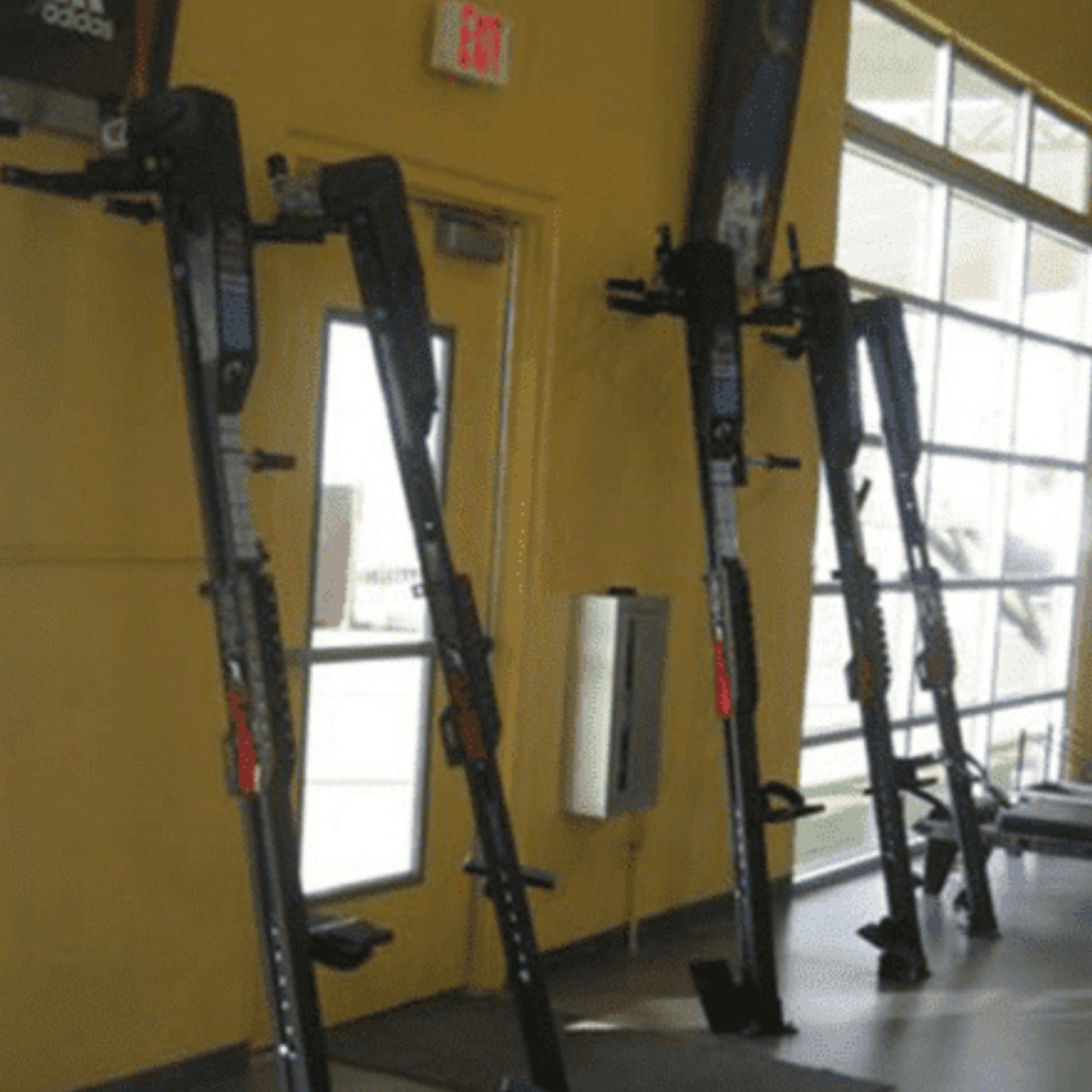 VersaClimber H/HP vertical climbing machines securely mounted on wall brackets in a commercial gym setting, displayed against a yellow wall with natural light from windows, highlighting professional installation