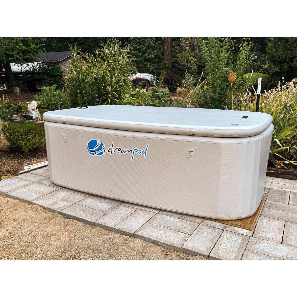 Dreampod rectangular ice bath with beige exterior and logo, installed on a paved patio surrounded by trees and garden greenery