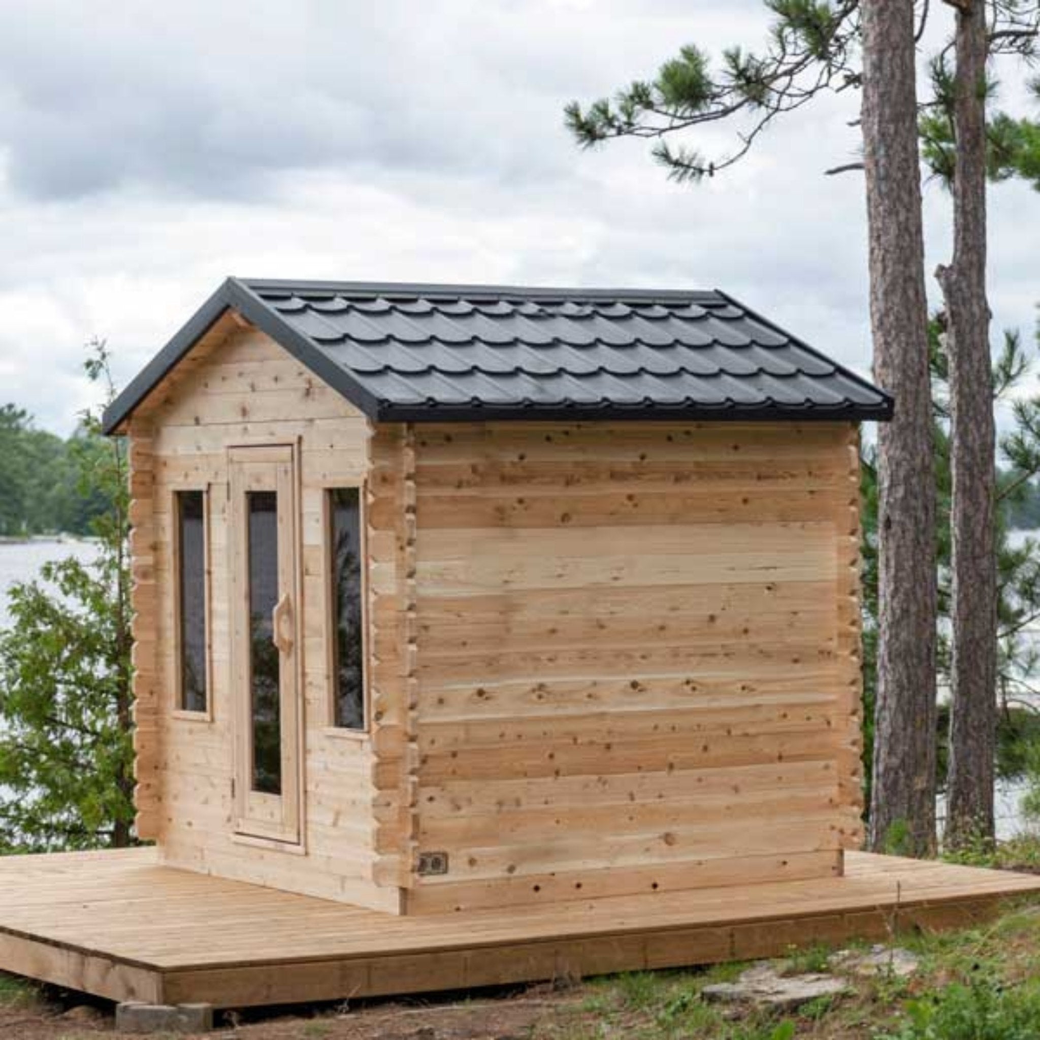 Leisurecraft CT Georgian Cabin Sauna with black shingled roof, cedar walls, and front deck, situated in a forest setting beside the water.