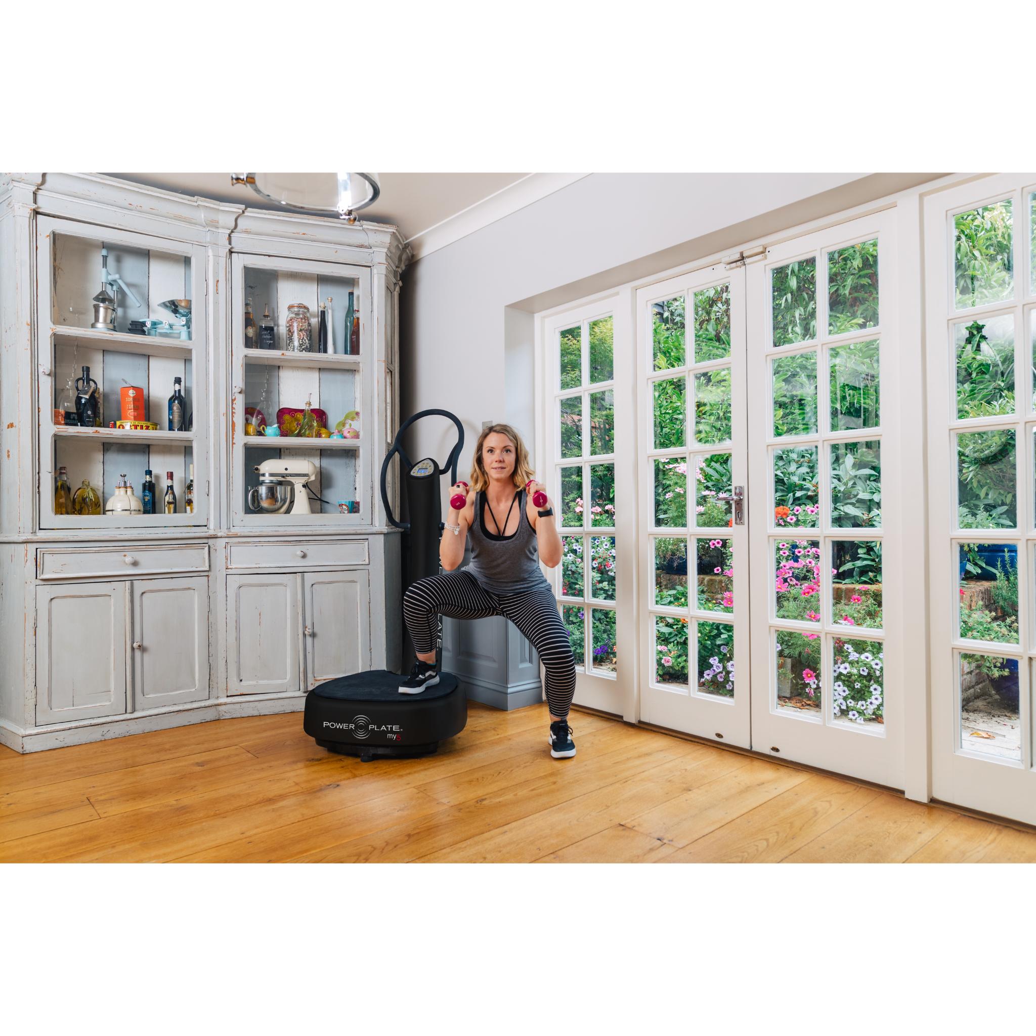 Woman exercising on a black Power Plate my5 vibration plate with straight handlebars, holding weights in a bright home setting near large glass doors