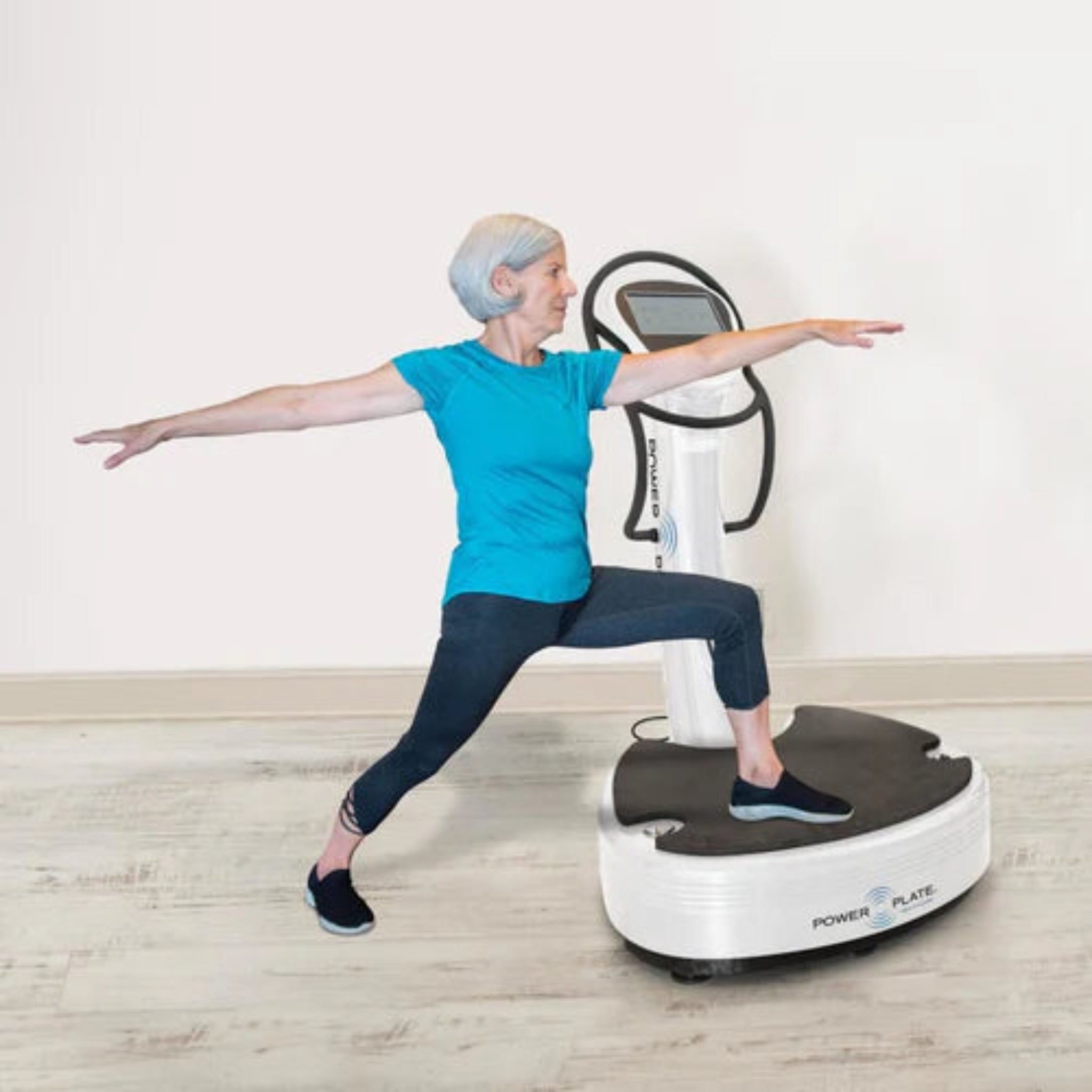Senior woman performing a balance exercise on the Power Plate pro7HC white vibration training machine in a home setting