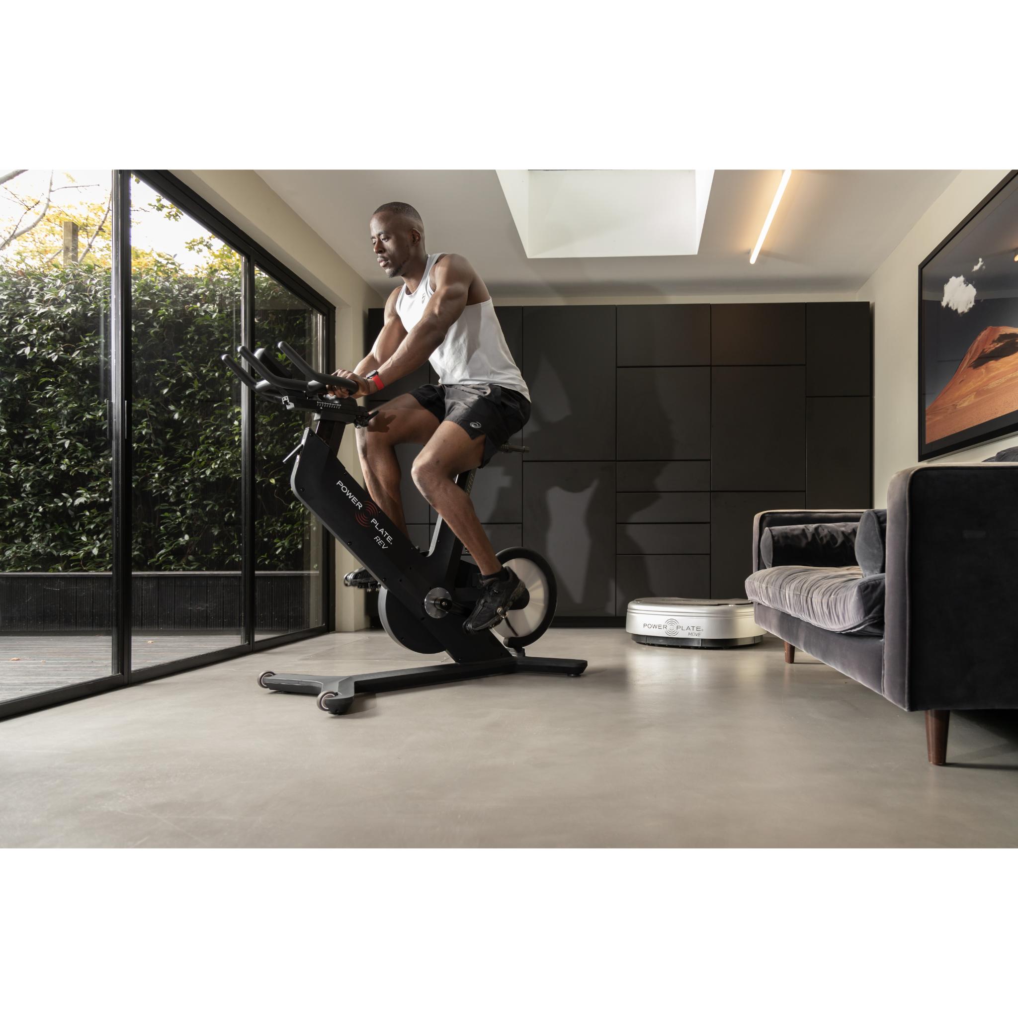Man exercising on the Power Plate REV black vibration training bike in a modern home gym setting with natural light