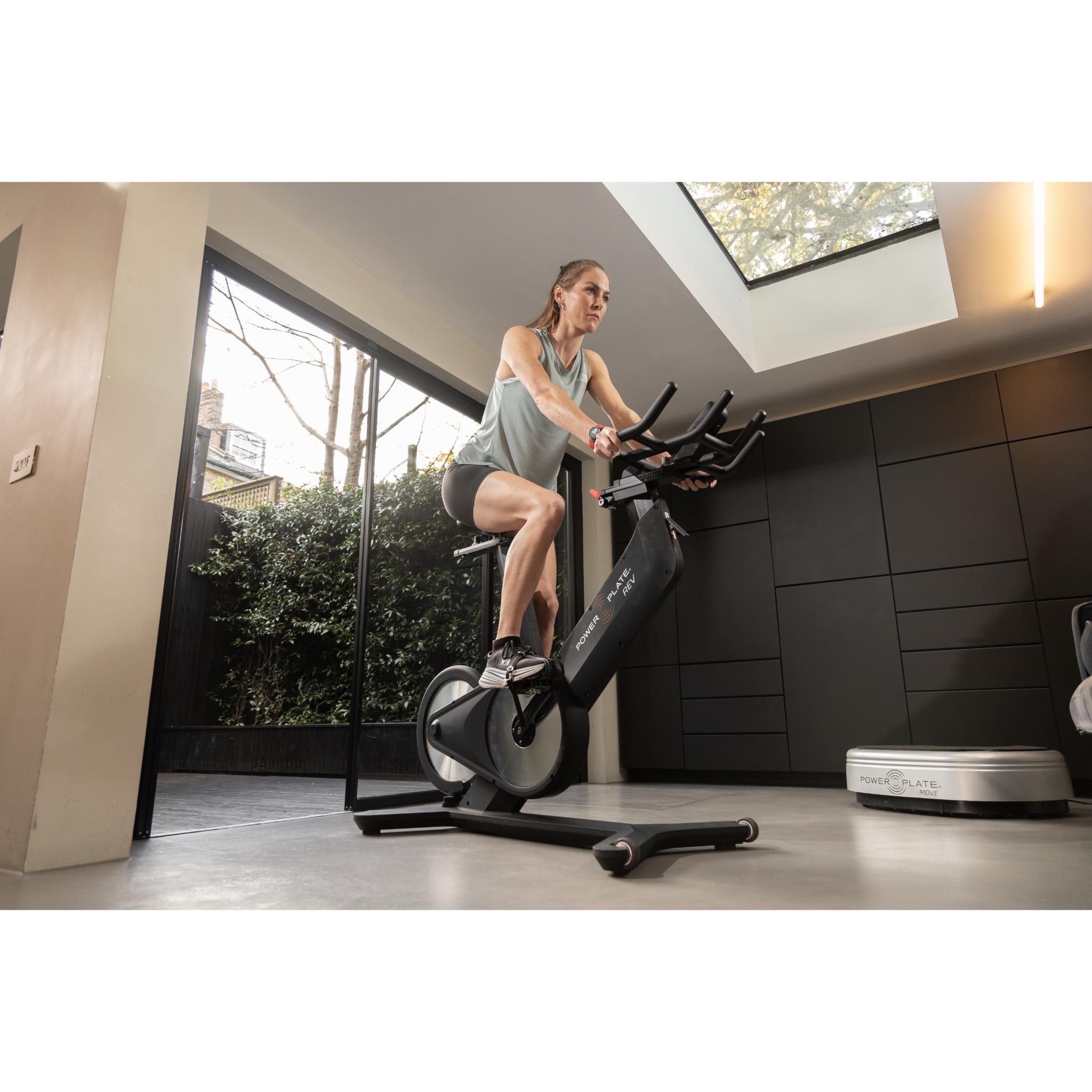 Woman exercising on the Power Plate REV black vibration training bike in a modern home gym with natural light