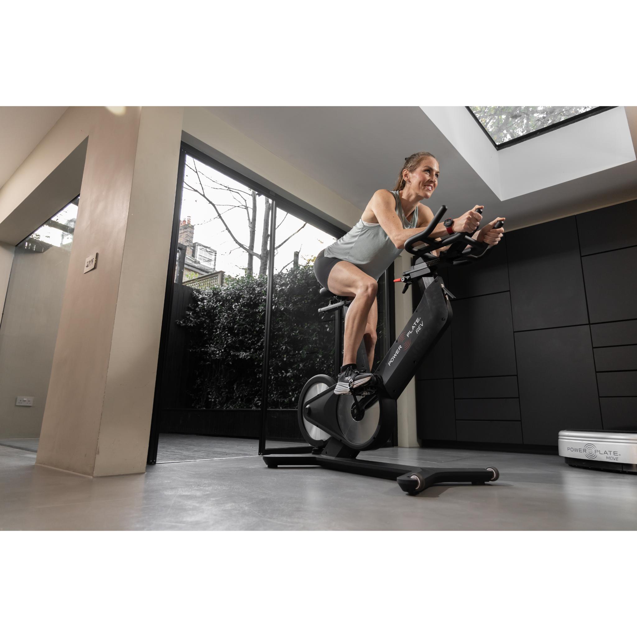 Woman exercising on the Power Plate REV black vibration training bike in a modern home gym with natural light