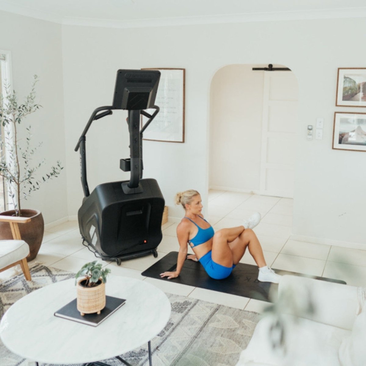 Woman stretching on a yoga mat in a living room with the STEPR Classic stair climber for home fitness.