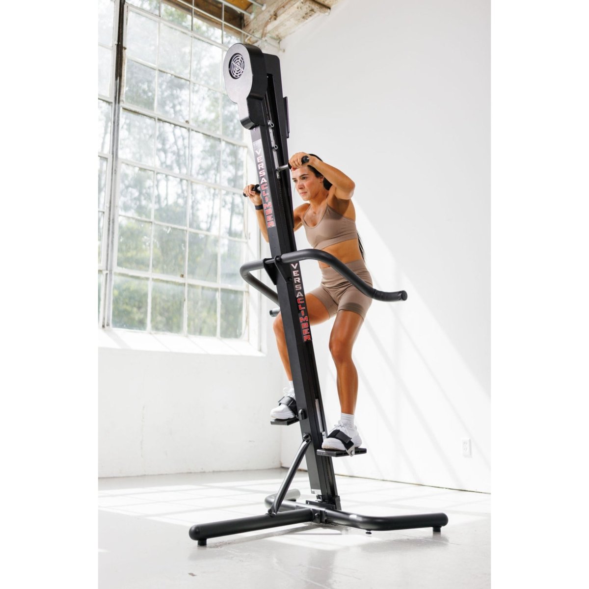Woman exercising on the VersaClimber TS-M Magnetic in regular black, performing full-body zero-impact cardio and strength training in a bright studio with large windows.